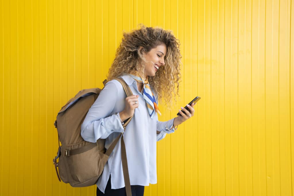 Smiling young woman with backpack in front of yellow background looking at cell phone